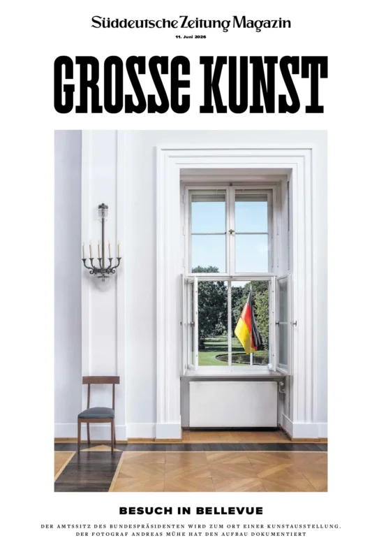Ein großes Fenster in einem hellen Raum bietet einen Blick auf einen grünen Rasen, auf dem draußen eine deutsche Flagge weht; im Raum sind zwei schwarze Stühle und eine Kerzenleuchte zu sehen.